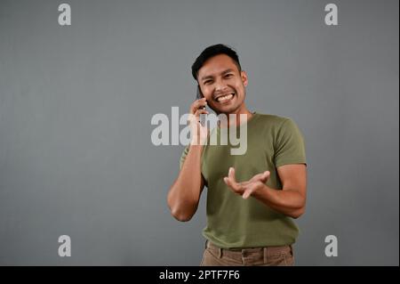 Allegro uomo asiatico millenario in abiti casual ama parlare al telefono con qualcuno mentre si trova su uno sfondo grigio isolato. Foto Stock