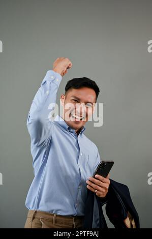 Gioioso uomo d'affari asiatico millenario alzando la mano, gioendo, celebrando il suo successo, tenendo la sua tuta e smartphone mentre si è in piedi su isolato Foto Stock