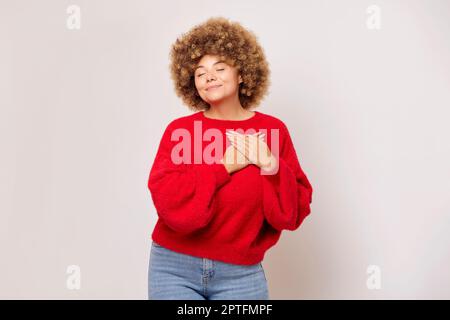 Cute giovane donna con capelli afro stile indossando maglione rosso si alza su sfondo chiaro, è felice e sorride allegro con gli occhi chiusi premendo han Foto Stock