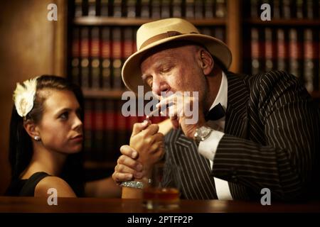 HES in controllo di tutti intorno a lui. Vecchio capo mob indossare un cappello e guardare serio, mentre una donna accende una sigaretta per lui Foto Stock