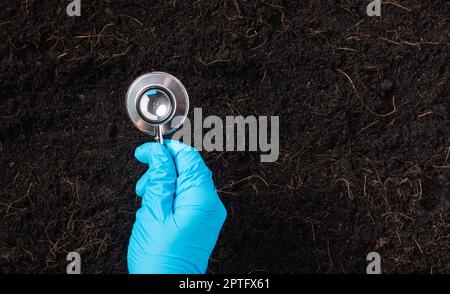 Mano di donna ricercatrice indossare guanti che tengono uno stetoscopio su terreno fertile nero per controllare la condizione prima di agricoltura o piantare, concetto di mondo Foto Stock