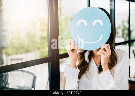 Revisione dell'esperienza del cliente. Donna che tiene sorridere emoji, donna felice di affari mostra la valutazione eccellente con l'icona del volto sorridente su carta circolare blu in negozio Foto Stock