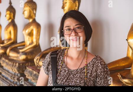 Ritratto di una donna turistica asiatica in un tempio della Thailandia, donna di mezza età. indossa occhiali da vista, sorridendo e guardando la fotocamera, sfondo sfocato o. Foto Stock