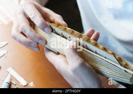 Mani di uomo incollando con colla i particolari del compensato per modello di nave, tenendo con le dita. Processo di costruzione di navi giocattolo, hobby, artigianato. Tavolo con variou Foto Stock