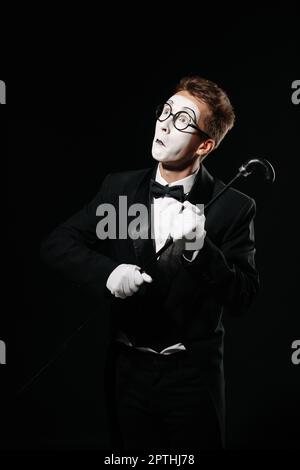 Ritratto di uomo mime in smoking e bicchieri in posa con bastone su sfondo nero Foto Stock