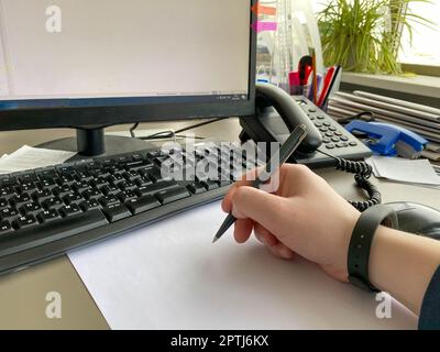 La mano di un uomo in una camicia e con un braccialetto per il fitness tiene una penna e scrive sul tavolo al tavolo dell'ufficio con un computer con una tastiera. Business Foto Stock