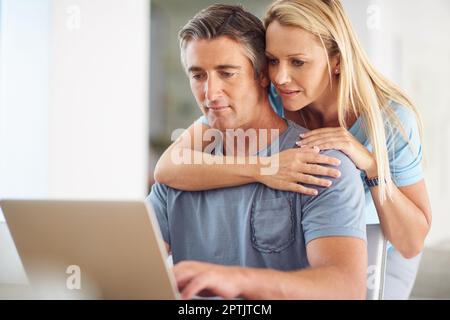 Cosa ne pensi? una donna matura e attraente che abbraccia il suo bel marito maturo mentre usa un computer portatile in casa Foto Stock