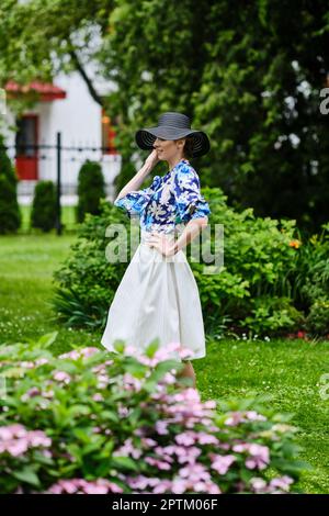 Ritratto di ragazza sorridente in cappello nero largo, blusa blu e gonna bianca in piedi sull'erba nel parco. Foto Stock