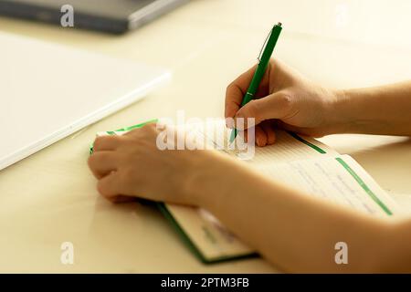 mani di una giovane donna bianca, ufficio a casa, lavora in remoto e studia online, prende appunti in un notebook con una penna a sfera verde su una scrivania bianca Foto Stock