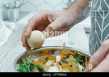 Mani maschili che sbucciano il guscio dell'uovo bollito sopra la ciotola di bucce vegetali primo piano. Cuocete sul tavolo da cucina, preparate gli ingredienti, gli scarti di cibo. Noce sana Foto Stock