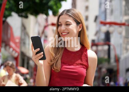 Ritratto di bella donna sorridente a piedi a San Paolo City SMS su telefono cellulare con sfondo sfocato Foto Stock