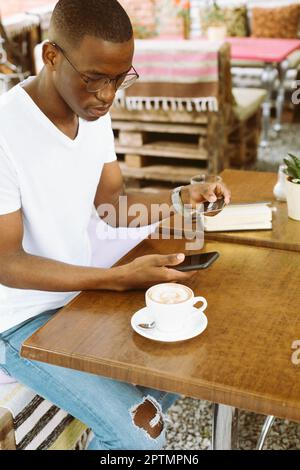 Uomo afro-americano con bicchieri seduti al tavolo in caffetteria con tazza di caffè sulla veranda estiva, tenendo smartphone e inserendo il numero di carta di credito Foto Stock