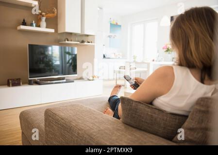 Donna incinta seduta sul divano e guardando la TV con telecomando, Monaco, Baviera, Germania Foto Stock