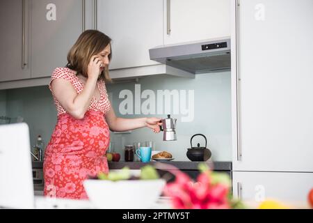 Donna incinta che parla su smartphone e fare il caffè per la colazione in cucina, Monaco, Baviera, Germania Foto Stock
