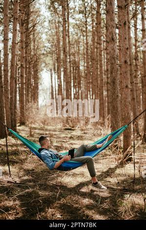 Giovane uomo che si rilassa in amaca nella foresta e che lavora su un tablet digitale Foto Stock