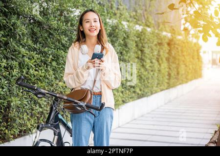 Giovane donna asiatica che si reca in bicicletta su smartphone in estate nel parco di campagna all'aperto, felice donna sorridente a piedi in strada con la sua bicicletta in città Foto Stock