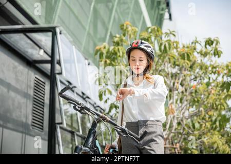 Ritratto sorridente donna controllare orologio, asiatico giovane donna d'affari in piedi con bicicletta controllare l'ora su edifici moderni città strada al mattino prima di andare Foto Stock