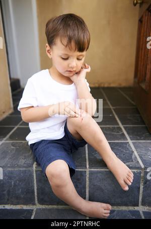 Anche i ragazzi più duri si feriscono. un ragazzino carino seduto su un gradino e guardando un taglio sul ginocchio Foto Stock