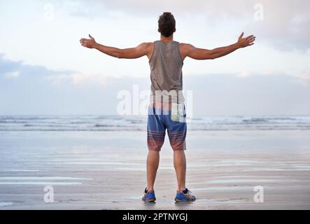 Abbracciando una bella mattina. Ripresa da dietro di un giovane uomo in piedi sulla spiaggia con le braccia sollevate verso il cielo Foto Stock