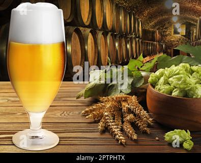 Bicchiere di gustosa birra leggera, luppolo fresco e punte di grano sul tavolo di legno in cantina Foto Stock