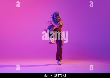 Una ragazza splendida, giovane ballerina moderna che fa trucchi hip-hop su sfondo viola studio in luce al neon. Break dance Street style Foto Stock