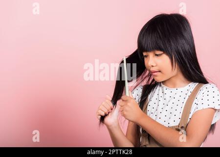 Bambino asiatico piccolo 10 anni tenere pettine spazzolando la sua unruly lei toccando i suoi lunghi capelli neri in studio shot isolato su sfondo rosa, bambino felice g Foto Stock