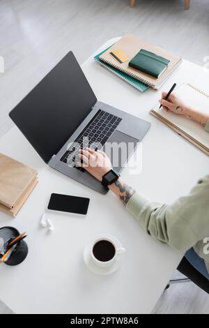 Vista dall'alto della scrittura del freelancer tatuata sul taccuino e usando il laptop vicino al caffè ed ai libri sul tavolo, immagine di scorta Foto Stock