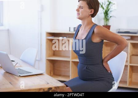 Soldato attraverso il dolore. Una donna d'affari incinta che lotta con mal di schiena alla sua scrivania ufficio Foto Stock