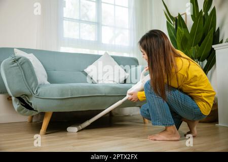 Casalinga donna polvere pulizia pavimento sotto divano o mobili divano con aspirapolvere, felice asiatico giovane donna con accumulatore aspirapolvere a hom Foto Stock