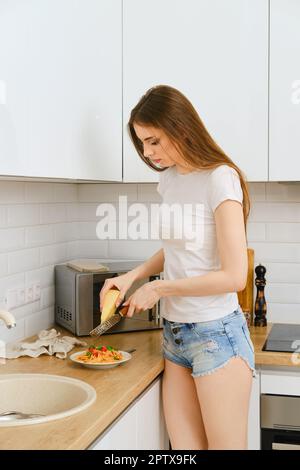 Bella giovane donna in t-shirt bianca e jeans corti grattugiano parmigiano su una grattugia a mano su un piatto di pasta in cucina Foto Stock