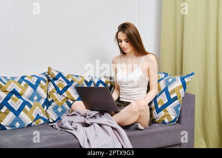 Giovane donna sorridente seduta sul divano di lavoro in remoto o di studio online su laptop a casa, freelance femminile che fa lavoro online a confortevole lavoro accogliente Foto Stock