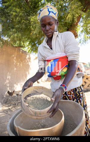 Una donna africana che porta il suo bambino sulla schiena usa un setaccio e calabash per raffinare la sua farina di grano miglio nella regione di Segou, Mali, Africa occidentale. 2022 siccità del Mali e crisi della fame. Foto Stock