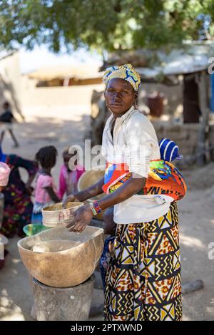 Una donna africana che porta il suo bambino sulla schiena usa un setaccio e calabash per raffinare la sua farina di grano miglio nella regione di Segou, Mali, Africa occidentale. 2022 siccità del Mali e crisi della fame. Foto Stock