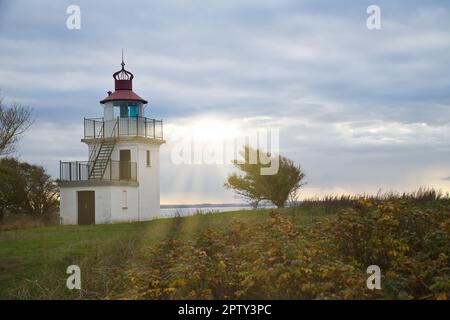 Faro, Spodsbjerg Fyr a Huntsted sulla costa della Danimarca. I raggi del sole brillano attraverso le nuvole. Prato con alberi. Foto di paesaggio dal mare Foto Stock