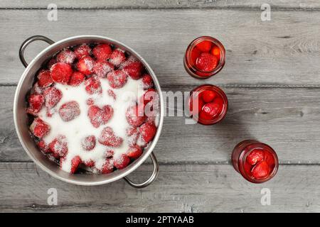 Vista del piano portapaziente, pentola in acciaio con fragole zucchero di cristallo sulla parte superiore, tre bottiglie con frutta sottaceto accanto ad essa. In casa Composta di fragola preparazione. Foto Stock