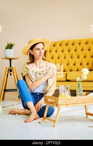 Ragazza a piedi nudi in jeans, camicia e cappello di paglia seduta sul pavimento vicino tavolo laterale con bevanda fresca Foto Stock