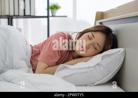 Giovane donna asiatica in pigiama rosa che dorme nel letto appoggiato sui cuscini e coprente piumino bianco. L'atmosfera della camera da letto nel tardo pomeriggio, su Foto Stock