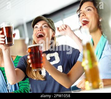 Grande schermo sportivo al bar. Tre amiche che si tirano indietro con qualche drink al bar e guardano lo sport Foto Stock