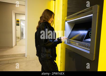 Giovane ragazza brunetta che ritira denaro dalla carta di credito al bancomat giallo. Foto Stock