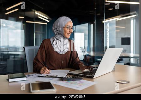 Giovane donna musulmana in programmatore hijab, sviluppatore che lavora in ufficio dietro notebook e con documenti. Foto Stock