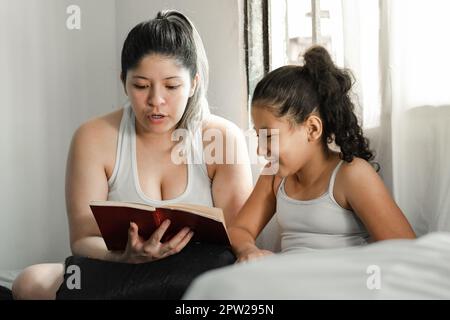 ragazza marrone sorridente mentre sua madre le legge un libro, seduta sopra il letto Foto Stock