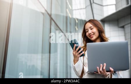 Donna d'affari sicura che utilizza smartphone con computer portatile in piedi in città. Bella donna d'affari pronta a lavorare. Concetto aziendale. Foto Stock