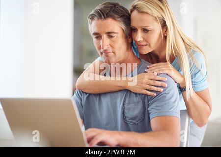 Cosa ne pensi? una donna matura e attraente che abbraccia il suo bel marito maturo mentre usa un computer portatile in casa Foto Stock