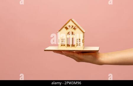 La mano di una donna tiene un modello in miniatura di una casa, un mock-up di un cottage su uno sfondo rosa. Acquisto di immobili, nuovi immobili, proprietà, contro Foto Stock