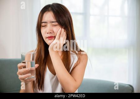 Bella femmina mal di denti e problemi dentali toccando guancia sensazione di dolore dopo aver bevuto acqua fredda, giovane donna asiatica con denti sensibili tenendo g Foto Stock