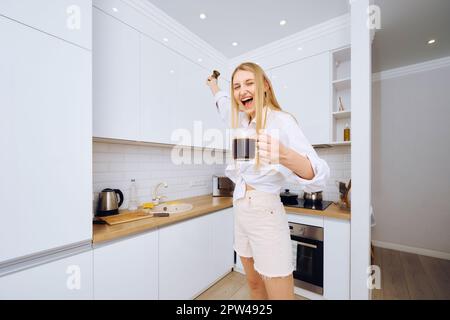 Donna allegra che balla e canta con una tazza di caffè in mano in cucina Foto Stock