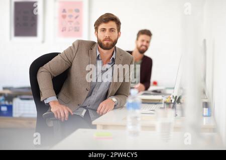 Prendendo sul serio il suo futuro di carriera. Serio giovane uomo d'affari seduto alla sua scrivania con un collega in background Foto Stock