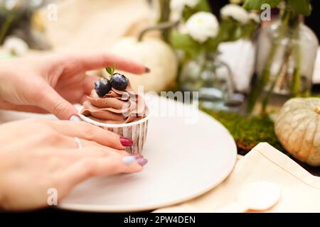 Delizioso cupcake con crema al cioccolato e mirtilli posti sul piatto vicino al bicchiere di vino e decorazioni biologiche sfocate Foto Stock