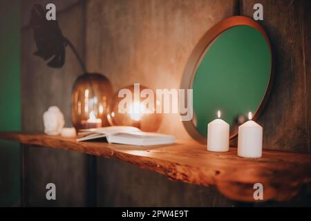 Specchio rotondo su ripiano di legno con due candele bruciate e una piccola testa di Buddha in porcellana riflessa in un angolo della superficie specchiata sopra la parete di cemento Foto Stock