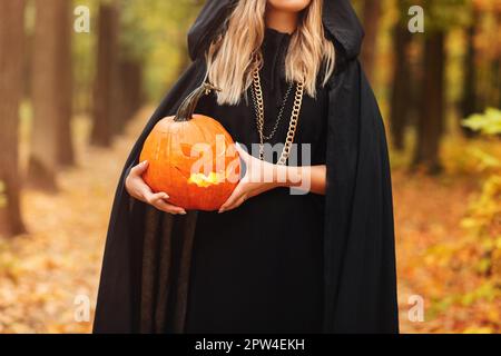 Giovane donna in mantello nero con jack o lanterna spooky E guardando via il giorno di Halloween nella foresta d'autunno Foto Stock